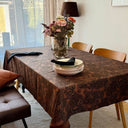 Närbild på tyget Astrid som bordsduk, med vävd struktur och dekorativt bladmönster i bruna och jordnära toner, kombinerat med dukning och blomsterarrangemang.