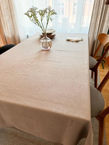Matbord täckt med en beige bordsduk med diskret vävd struktur, dukat med tallrikar, servetter och en glasvas med vita blommor framför ett ljust fönster.