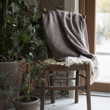 Beige Franke bomullspläd med offwhite fransar draperad över en trästol vid ett fönster