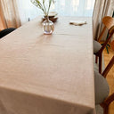 Matbord täckt med en beige bordsduk med diskret vävd struktur, dukat med tallrikar, servetter och en glasvas med vita blommor framför ett ljust fönster.