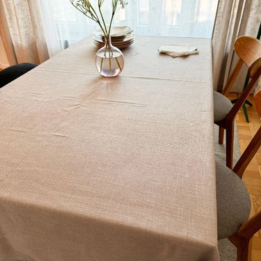 Matbord täckt med en beige bordsduk med diskret vävd struktur, dukat med tallrikar, servetter och en glasvas med vita blommor framför ett ljust fönster.
