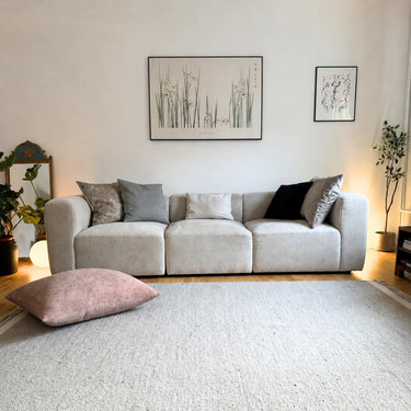 Puderrosa Milena sittkudde i möbeltyg placerad på golvet framför en ljus soffa.