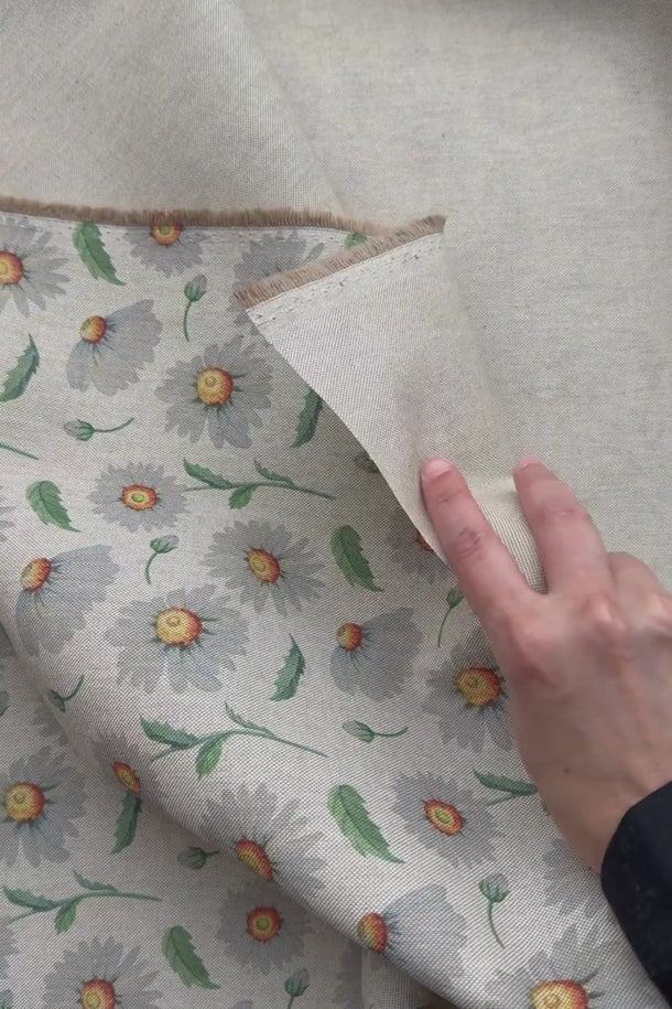 En hand visar och känner på tyget Daisy med prästkragemönster och linnelook för att demonstrera struktur, fall och materialkänsla.